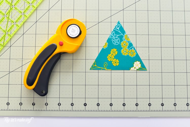 A fabric cutter, an Omnigrip ruler, and a piece of material cut into a triangle all sitting on a fabric mat