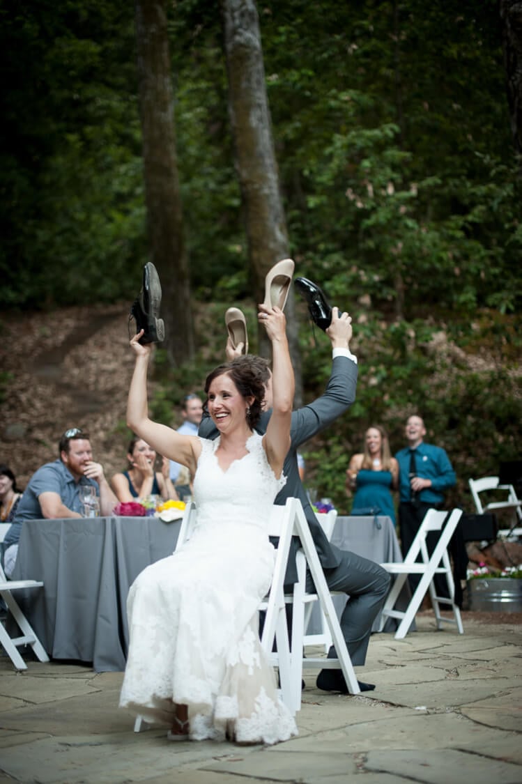 A bride and a groom sitting back-to-back and each holding one of each other\'s shoes in the air