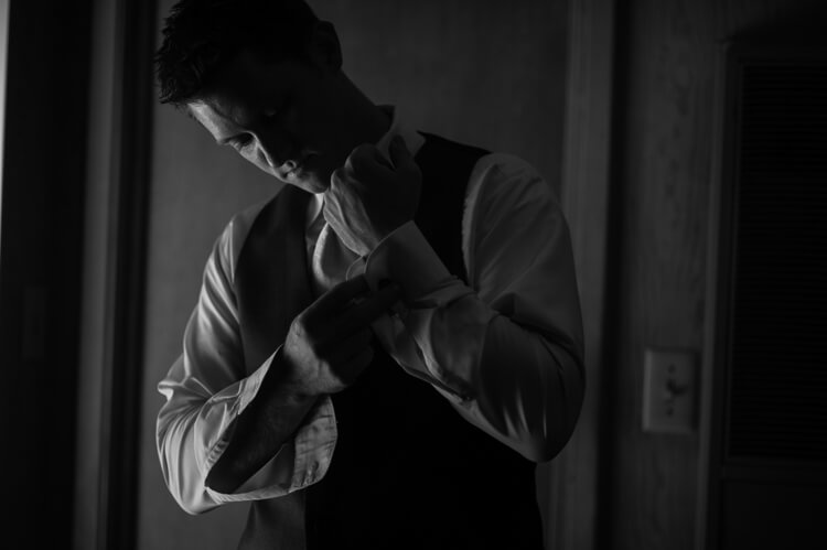 A man standing in a dark room putting on cufflinks on his long-sleeved shirt