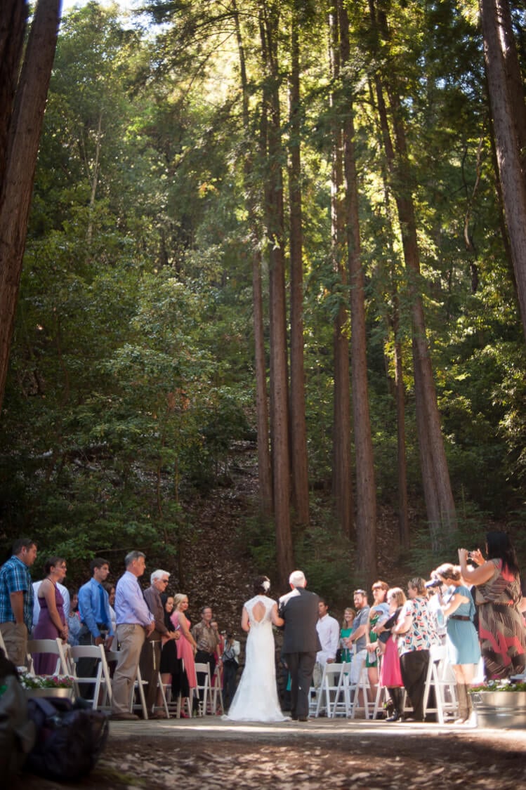 A group of people in the woods standing by chairs and watching the bride and father of the bride