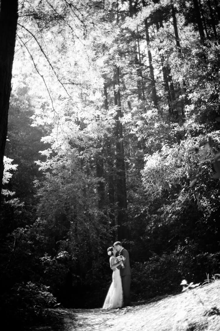 A black and white photo of a groom kissing his bride among tall trees