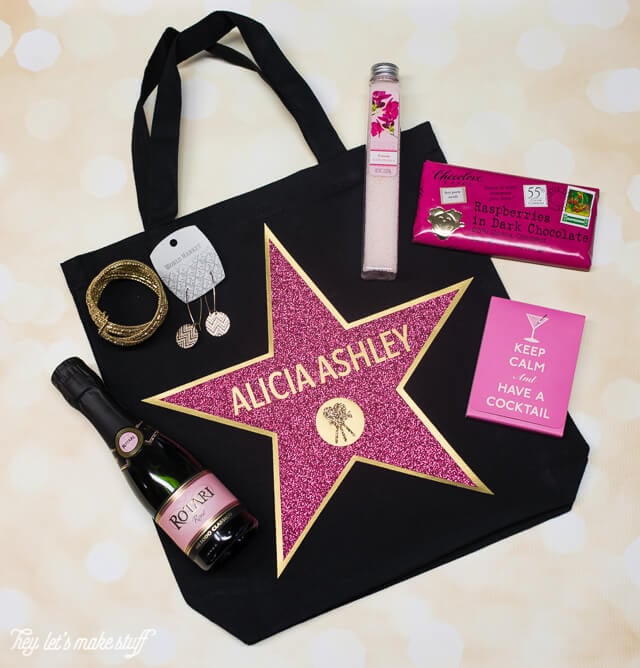 A black tote bag with an image of a gold star with a pink personalized star over the top of it and an image of a movie camera applied to it along with jewelry, champagne and other party favors on top of the bag