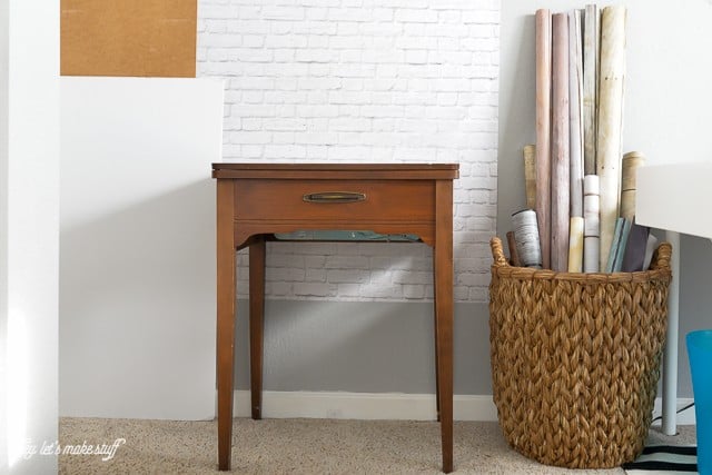 A small sewing table up against a white brick wall and a basket on the floor filled with rolls of paper