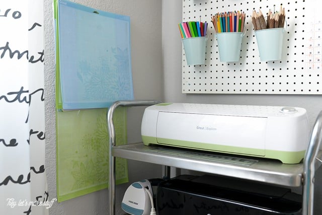 A peg board with markers in little buckets, a Cricut machine on a cart and Cricut mats hanging from a hook on the wall