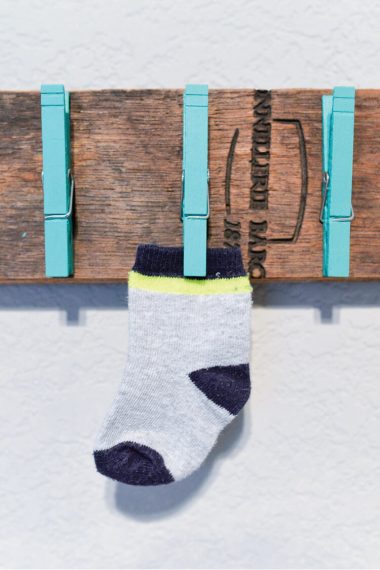 A piece of board hanging on a wall with three aqua colored clothespins attached to it and one clothespin has a sock clipped to it