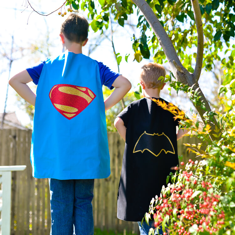 Two boys standing outside with their backs to the camera, and they are wearing a Superman and a Batman cape
