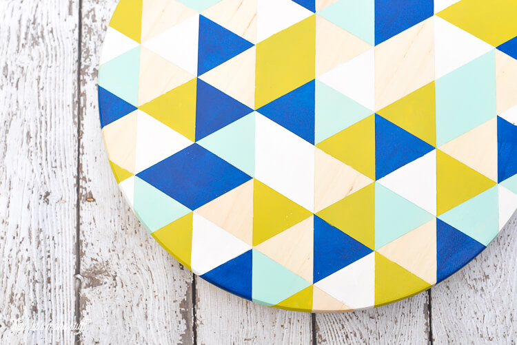 A round piece of wood with white, yellow, light blue, dark blue and peach painted triangles and diamonds, sitting on a wooden table