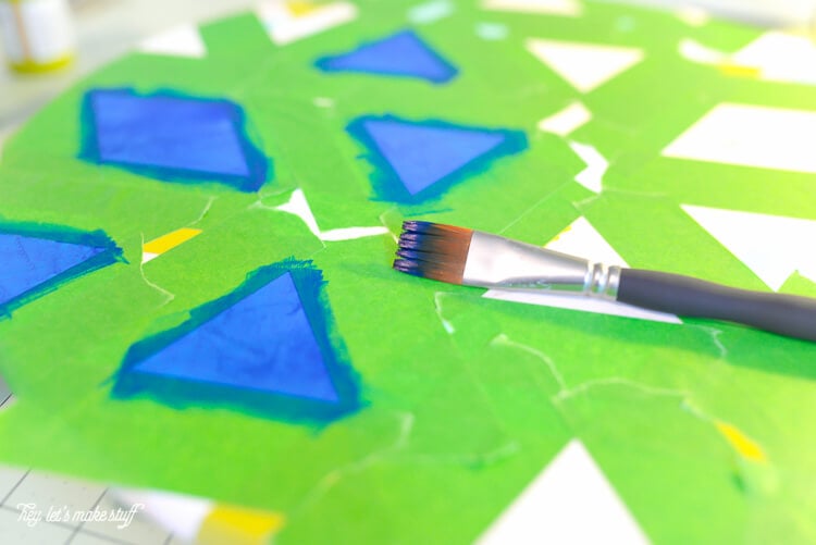 A paint brush and a round piece of wood with green tape all over it with dark blue painted triangles and diamonds