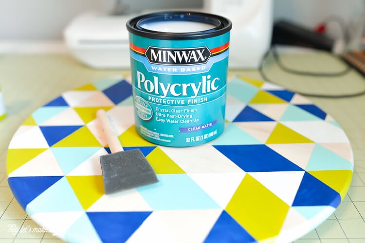 A can of Minwax polycrylic protective finish on top of a round piece of painted wood and a sponge paintbrush