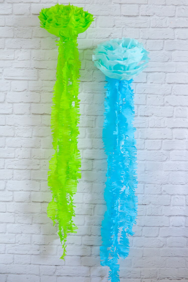 Green and blue tissue paper hanging on a white brick wall.  The tissue paper resembles jellyfish