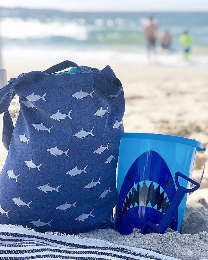A blanket, a blue tote bag with sharks on it, a blue plastic bucket decorated with a shark image and a blue plastic sand scooper all sitting on the beach