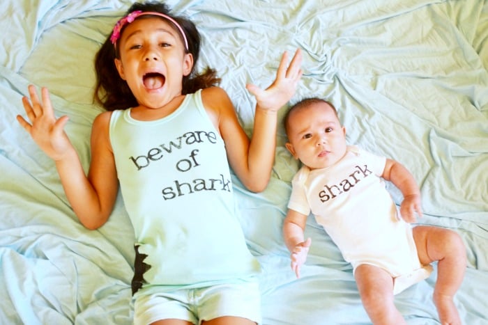A young girl and a baby lying on a bed with the girl wearing a tank top that says, \"Beware of Shark\" and the baby wearing a onesie that says, \"Shark\"