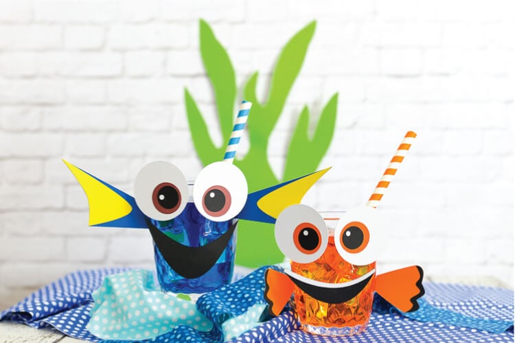 Two glasses with straws in them sitting on several pieces of blue fabric and the glasses are decorated to look like Dory and Nemo