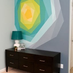 A dresser holding a lamp, a plant, a glass vase and some books with a wall mural on the wall behind it