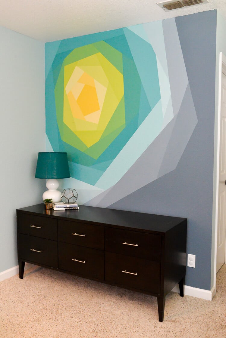 A dresser holding a lamp, a plant, a glass vase and some books with a wall mural on the wall behind it