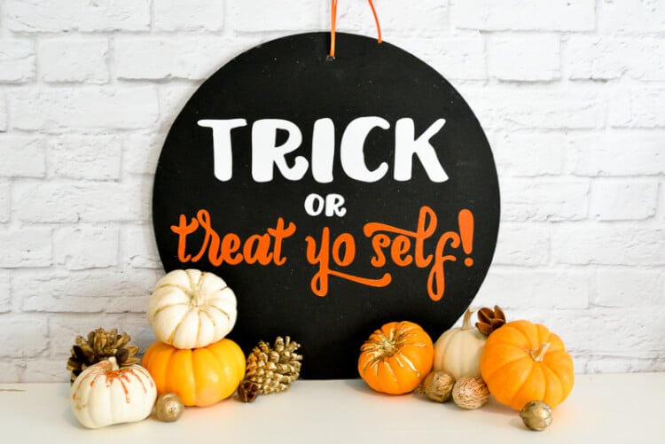 A round black sign sitting on a table and leaning up against a white brick wall.  The table is decorated with tiny pumpkins and the sign says, \"Trick or Treat yo self!\"