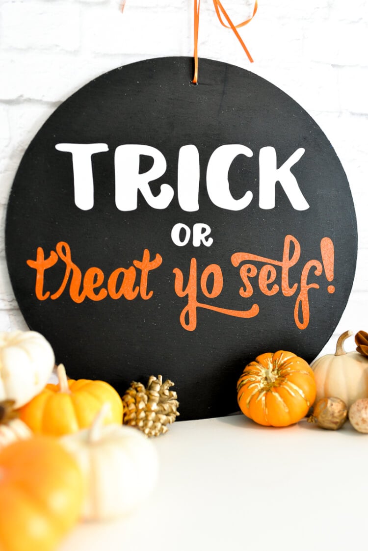Close up of a round black sign sitting on a table and leaning up against a white brick wall.  The table is decorated with tiny pumpkins and the sign says, \"Trick or Treat yo self!\"