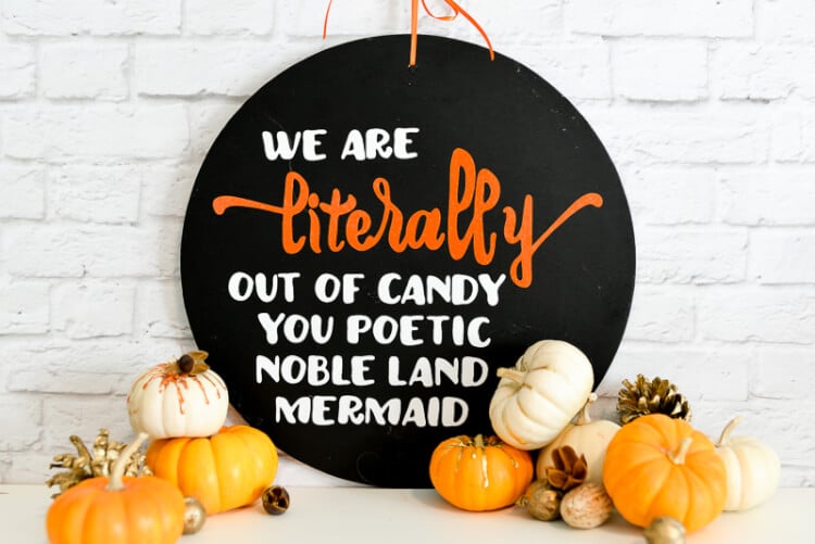 A round black sign sitting on a table and leaning up against a white brick wall.  The table is decorated with tiny pumpkins and the sign says, \"We are Literally Out of Candy You Poetic Noble Land Mermaid\"