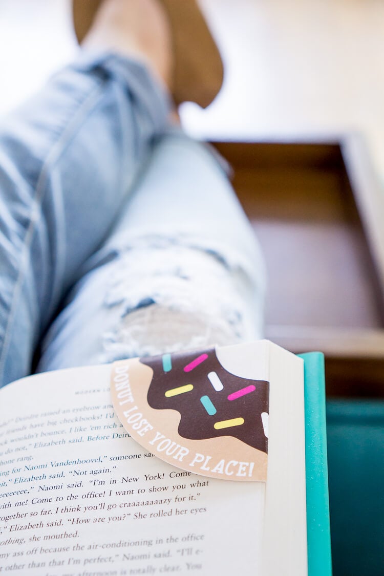 A person\'s legs propped up on a table holding an open book with a donut bookmark in the corner of a page