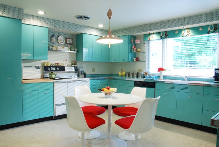 A kitchen pained in a teal color with a white table and chairs.  Chairs have a red cushion on them.