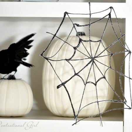 Black glitter spider web hanging in front of a white pumpkin sitting on a shelf