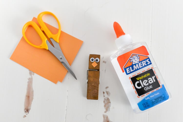 Elmer\'s glue next to a clothespin decorated with googly eyes, a pair of scissors and some orange paper