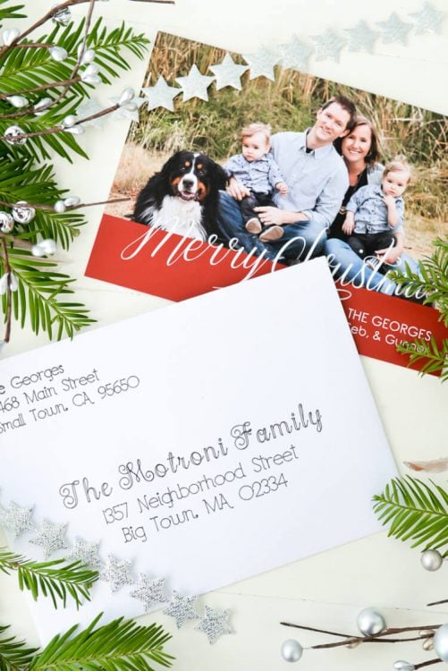 A Christmas card and an addressed envelope sitting next to some holiday greenery on a table