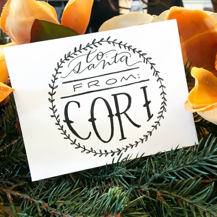 A white sign sitting on top of some yellow flowers and greenery that says, \"To Santa From Cori\"