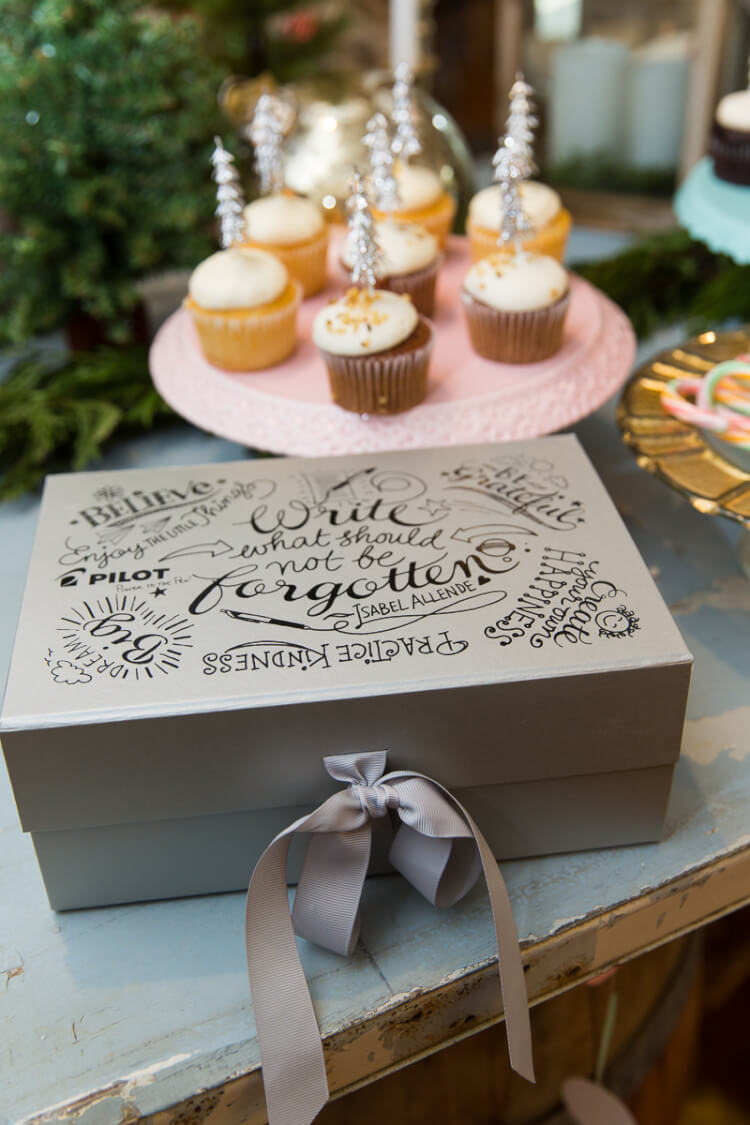 A plate of cupcakes and a box sitting on a table. The box is decorated with a bow and hand lettered flourishes