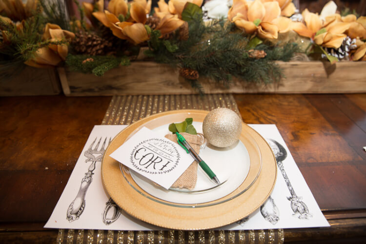 A place setting next to some flowers and the plate has a Christmas ornament on it and a pen with a hand lettered note