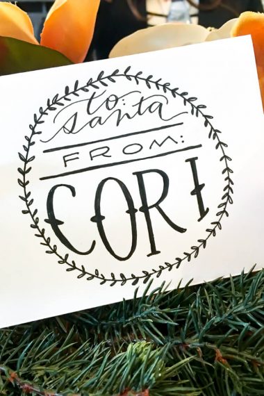 A white sign sitting on top of some yellow flowers and greenery that says, "To Santa From Cori"