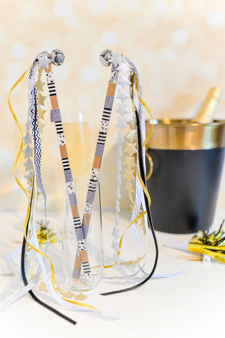 Sitting on a table are two champagne flutes, a bottle of champagne in an ice bucket and two party streamers with bells on them stuck inside a third champagne flute