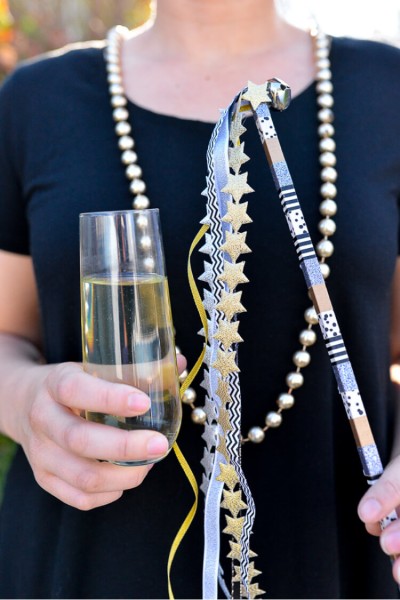 A person wearing a dark navy-blue dress and a string of pearls and holding a flute of champagne and a party streamer with bells
