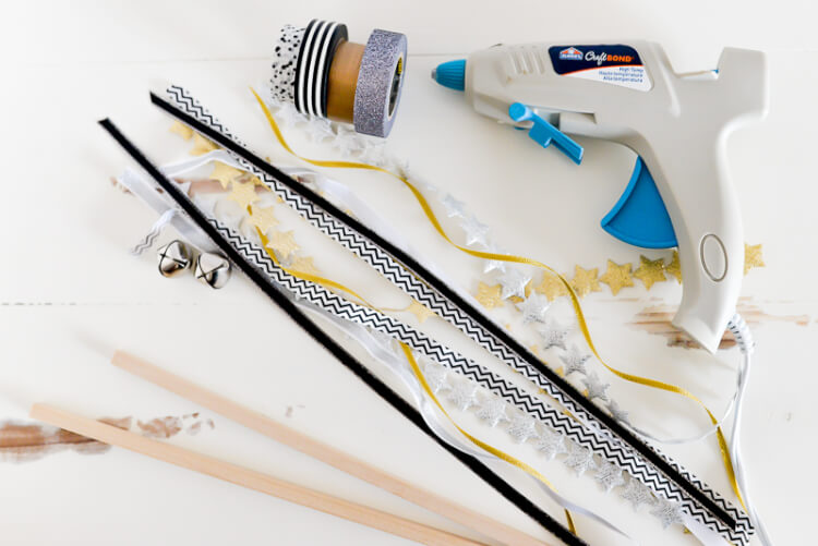 A hot glue gun, ribbon, small silver bells, star garlands and dowel sticks sitting on a table