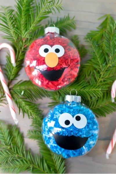 Some greenery, a candy cane and two glass ornaments decorated to look like Cookie Monster and Elmo from Sesame Street, all sitting on a table