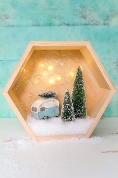 A wooden hexagon shaped shadow box that contains a retro camper with a small bottle brush tree on top of it and two other bottle brush trees standing next to the camper surrounded by fake snow and twinkle lights