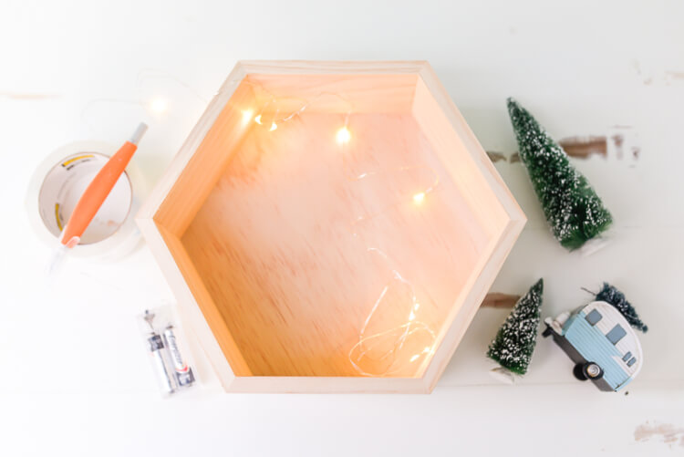 A wooden hexagon shaped shadow box with some twinkle lights in it and bottle brush trees and a retro camper and other crafting supplies all laying on a table