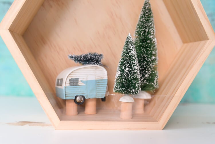 A close up of a wooden hexagon shaped shadow box that contains a retro camper with a small bottle brush tree on top of it and two other bottle brush trees standing next to the camper.  The camper and the two trees are sitting on top of wooden spools.