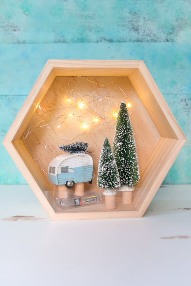 A close up of a wooden hexagon shaped shadow box that contains a retro camper with a small bottle brush tree on top of it and two other bottle brush trees standing next to the camper.  The camper and the two trees are sitting on top of wooden spools.