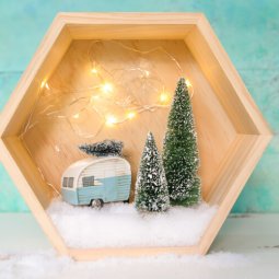 A close up of a wooden hexagon shaped shadow box that contains a retro camper with a small bottle brush tree on top of it and two other bottle brush trees standing next to the camper surrounded by fake snow and twinkle lights