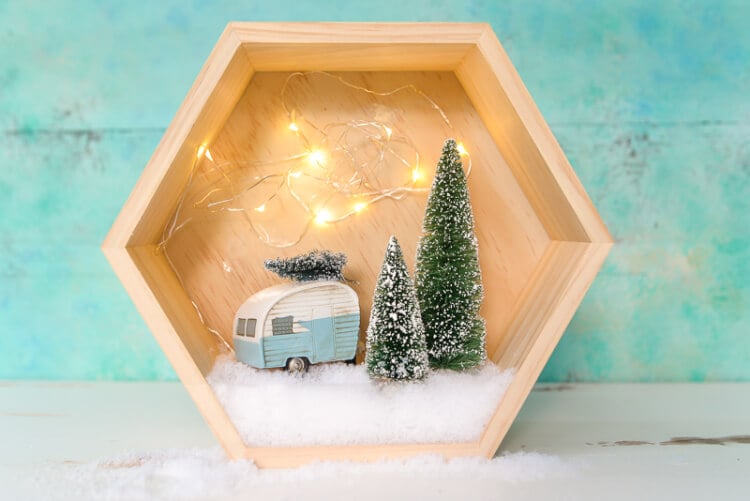 A close up of a wooden hexagon shaped shadow box that contains a retro camper with a small bottle brush tree on top of it and two other bottle brush trees standing next to the camper surrounded by fake snow and twinkle lights