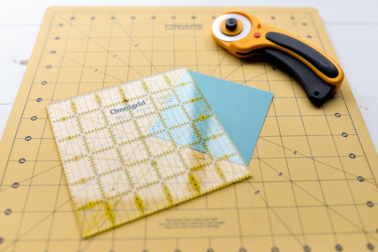 A ruler, a rotary cutter and a square piece of blue cardstock laying on top of a mat and a table