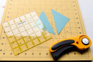 A ruler, a rotary cutter and a square piece of blue cardstock cut into two triangles laying on top of a mat and a table