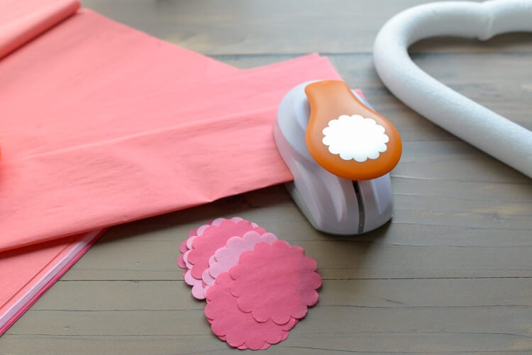 Heart-shaped foam wreath form, a scalloped-edge paper punch and pink tissue paper lying on a wooden table