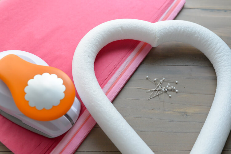 Heart-shaped foam wreath form, some straight pins, a scalloped-edge paper punch and pink tissue paper lying on a wooden table
