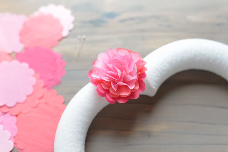 Sitting on a table are some tissue paper circles, some straight pins and a fluffed-up tissue paper circle that resembles a flower that is attached to a heart shaped wreath form