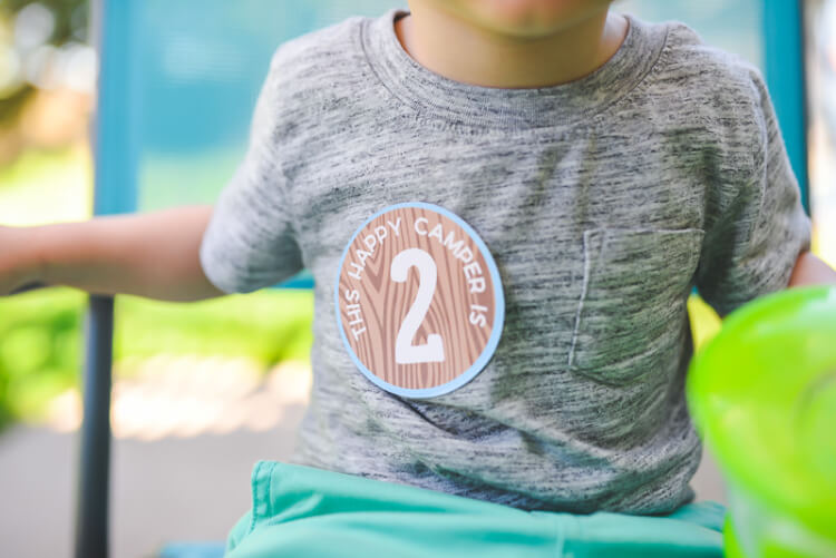 A close up of a little boy sitting on a chair, wearing aqua blue pants and a gray shirt that has a sticker on it.  The sticker says, \"This Happy Camper is 2\"
