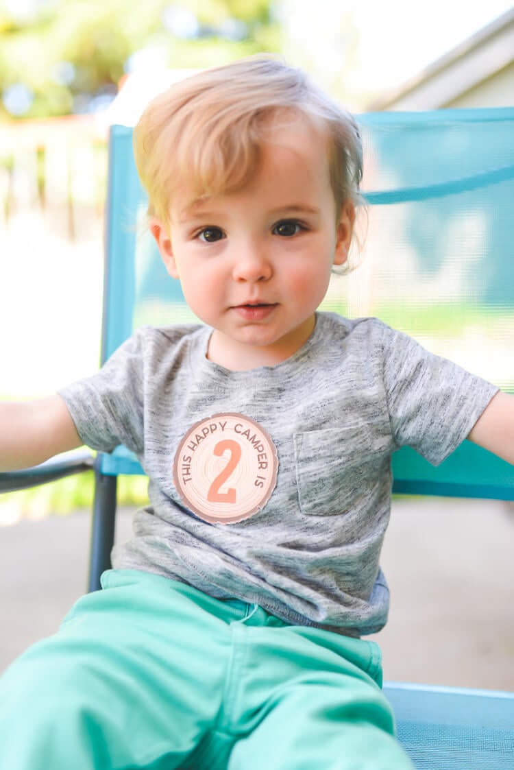 A little boy sitting on a chair, wearing aqua blue pants and a gray shirt that has a sticker on it.  The sticker says, \"This Happy Camper is 2\"