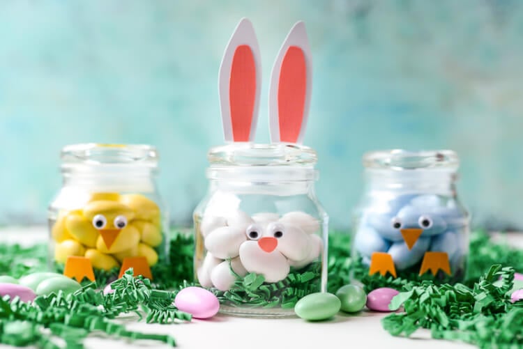 Three small jars on a table surrounded by greenery and Easter candy.  The jars are decorated to resemble a baby chick, a rabbit and a bird