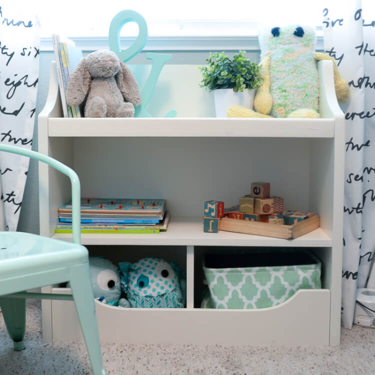 A book shelf next to a window, filled with books and toys and a mint green chair next to it
